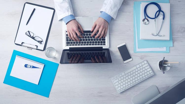 An overhead view of someone working on a laptop at a desk with paperwork, glasses and a stethoscope.